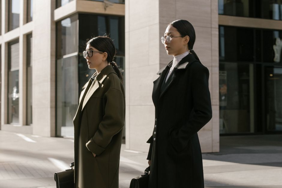 Two stylish women standing outside an urban office building, dressed in formal coats.
